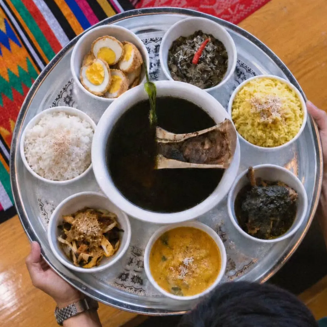 A Dulang tray seen from above: a bowl of black Tiyula Itum soup with bone marrow in the centre, surrounded by bowls of chicken pianggang, beef kurma, chicken belachan, kaliya puso, turmeric rice, white rice and eggs.