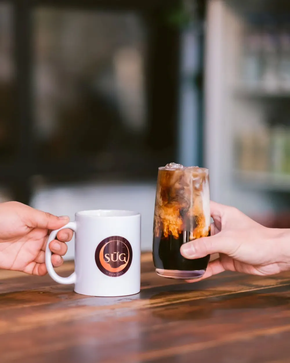 Two hands raising a hot mug and an iced glass of Sulu coffee.