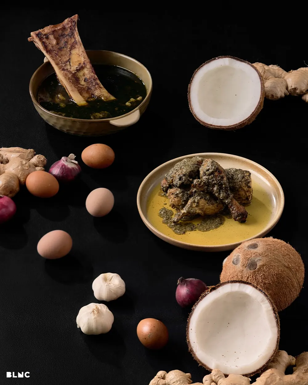 Coconut halves, ginger, eggs, shallots and garlic laid out on black around bowls of Tiyula Itum and pianggang.