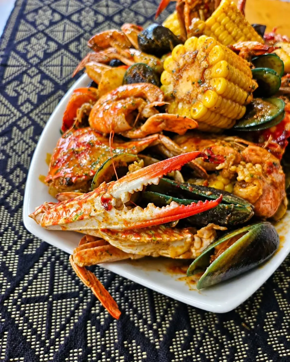 A platter of blue crabs, mussels, prawns and sweet corn in Montaño sauce on a dark woven cloth.